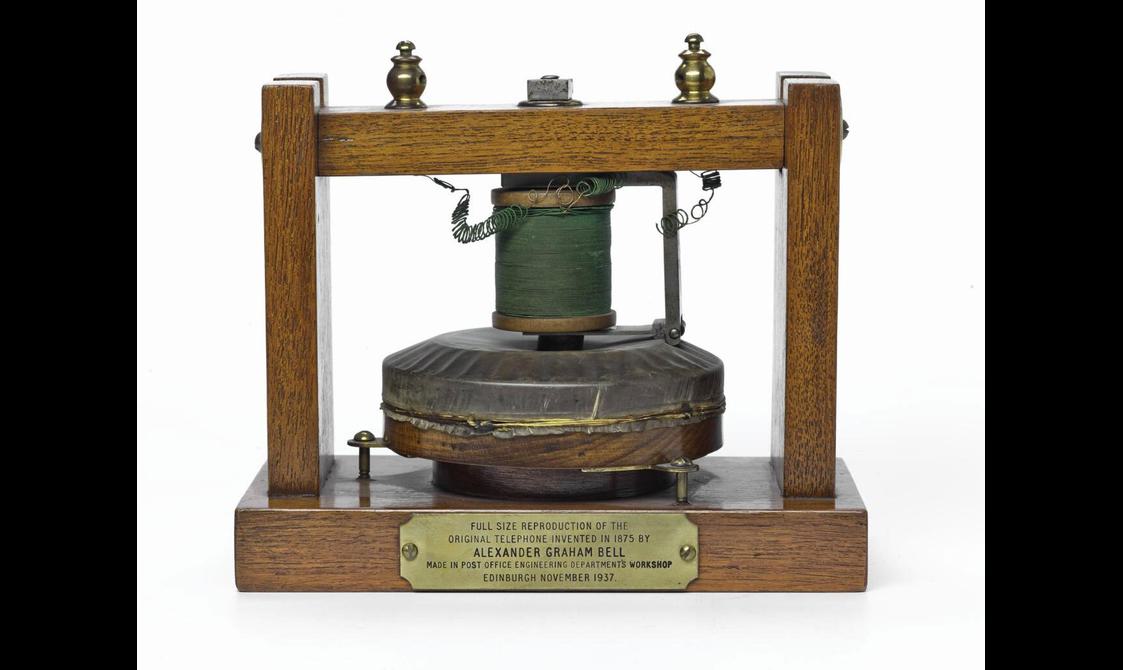An early telephone which has a wooden frame and a round object sitting on the base with a spool with wire on top.
