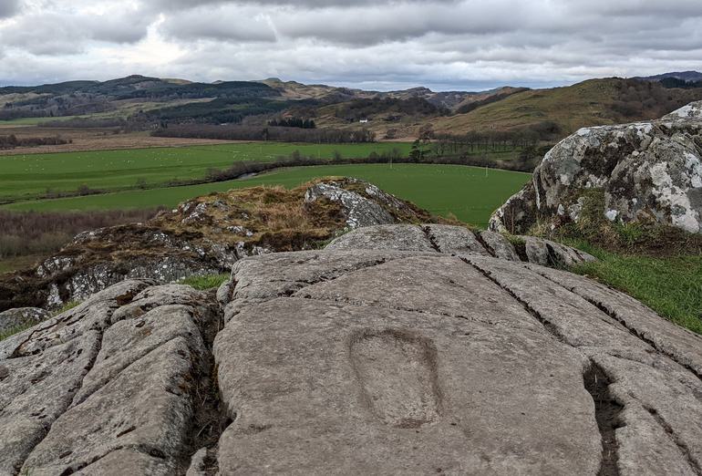 A rock with a footprint imprinted on its surface overlooking green fields and green and brown hills