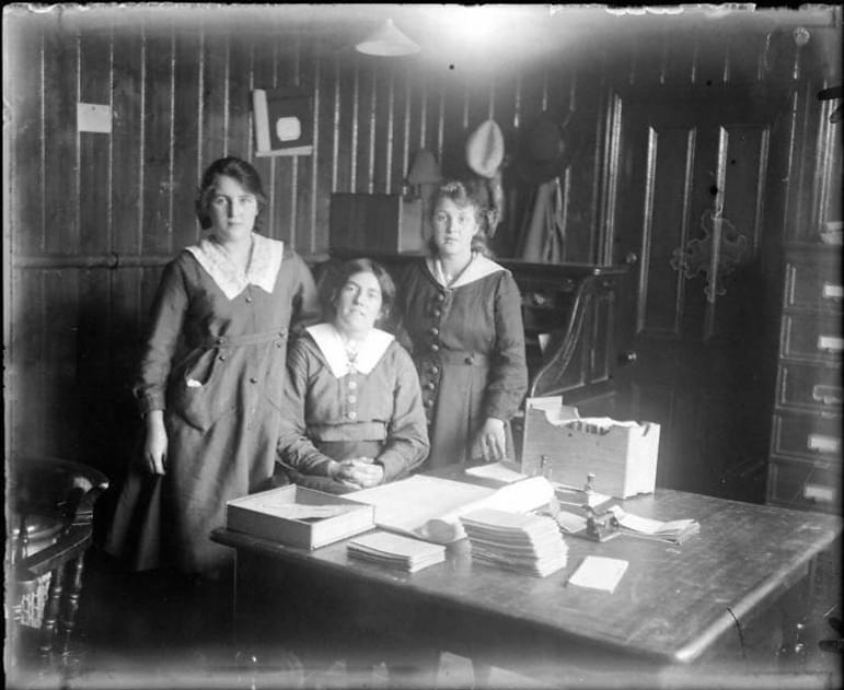 A black and white photograph of three clerical workers behind a desk in an early 20th century office.